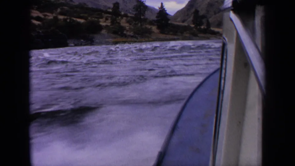 1963 Idaho 8mm frame from a moving boat, looking over blue-gray rapids toward trees, rocky hills, and distant mountains.
