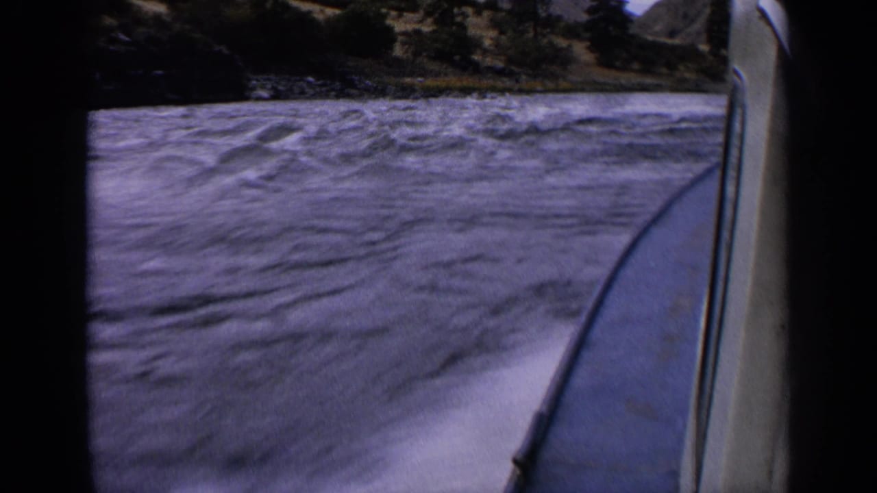 1963 Idaho home-movie frame showing rushing river water from a boat, with blurred spray, shoreline trees, and rocky terrain beyond.