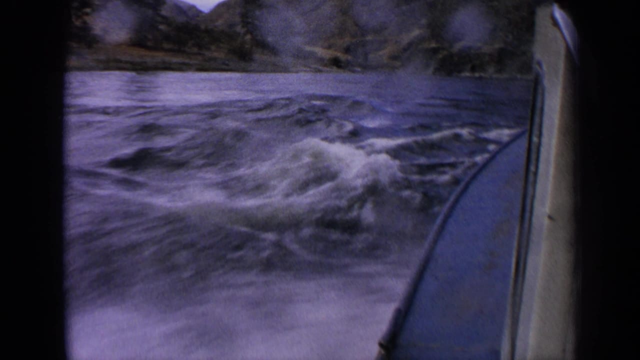 8mm home-movie frame showing a motorboat moving through foamy Idaho river water with rocky banks in the background.