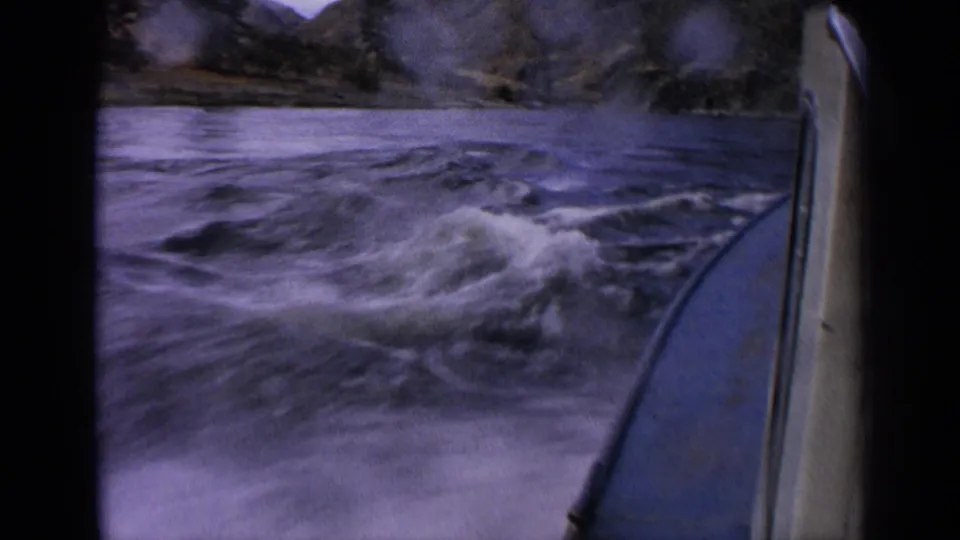 8mm home-movie frame showing a motorboat moving through foamy Idaho river water with rocky banks in the background.