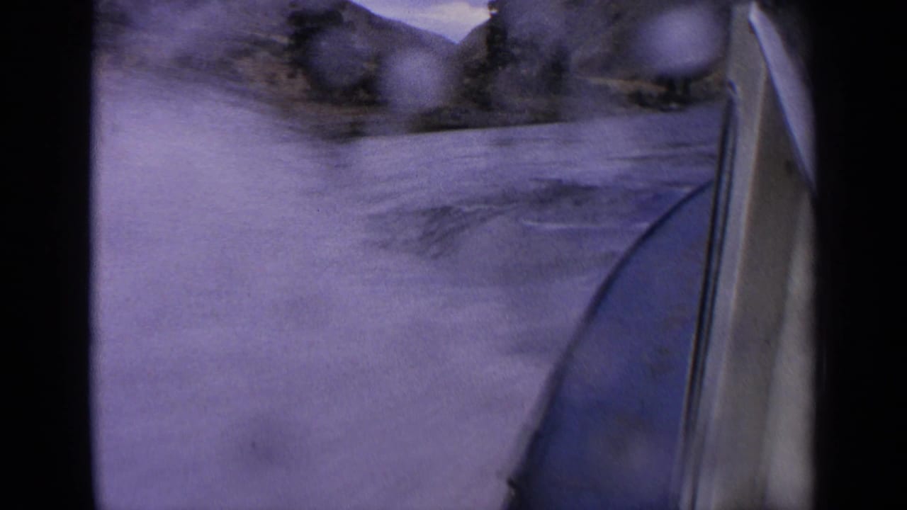 1963 Idaho 8mm frame from a moving boat, showing the bow beside rippled river water and rugged canyon slopes ahead.