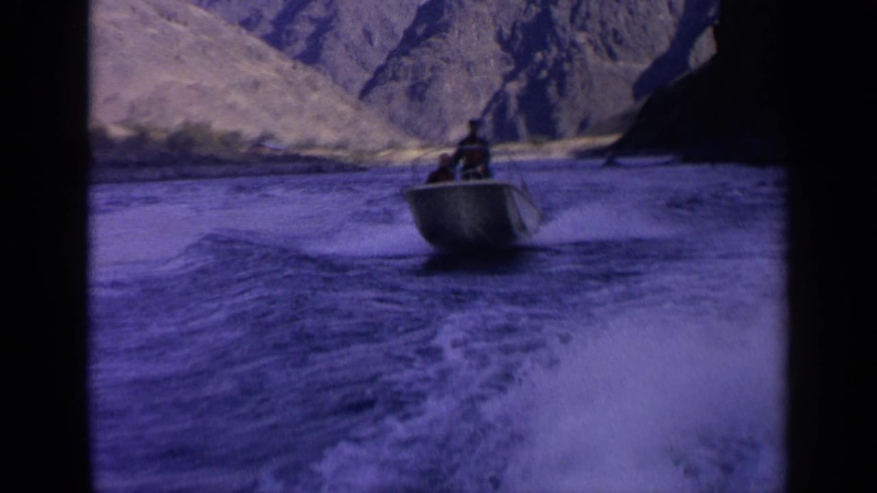 1963 Idaho home-movie frame showing a distant person in a small motorboat crossing foamy wake amid rugged canyon slopes.