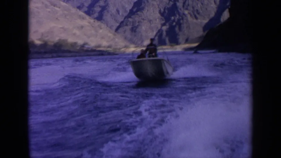 1963 Idaho home-movie frame showing a distant person in a small motorboat crossing foamy wake amid rugged canyon slopes.