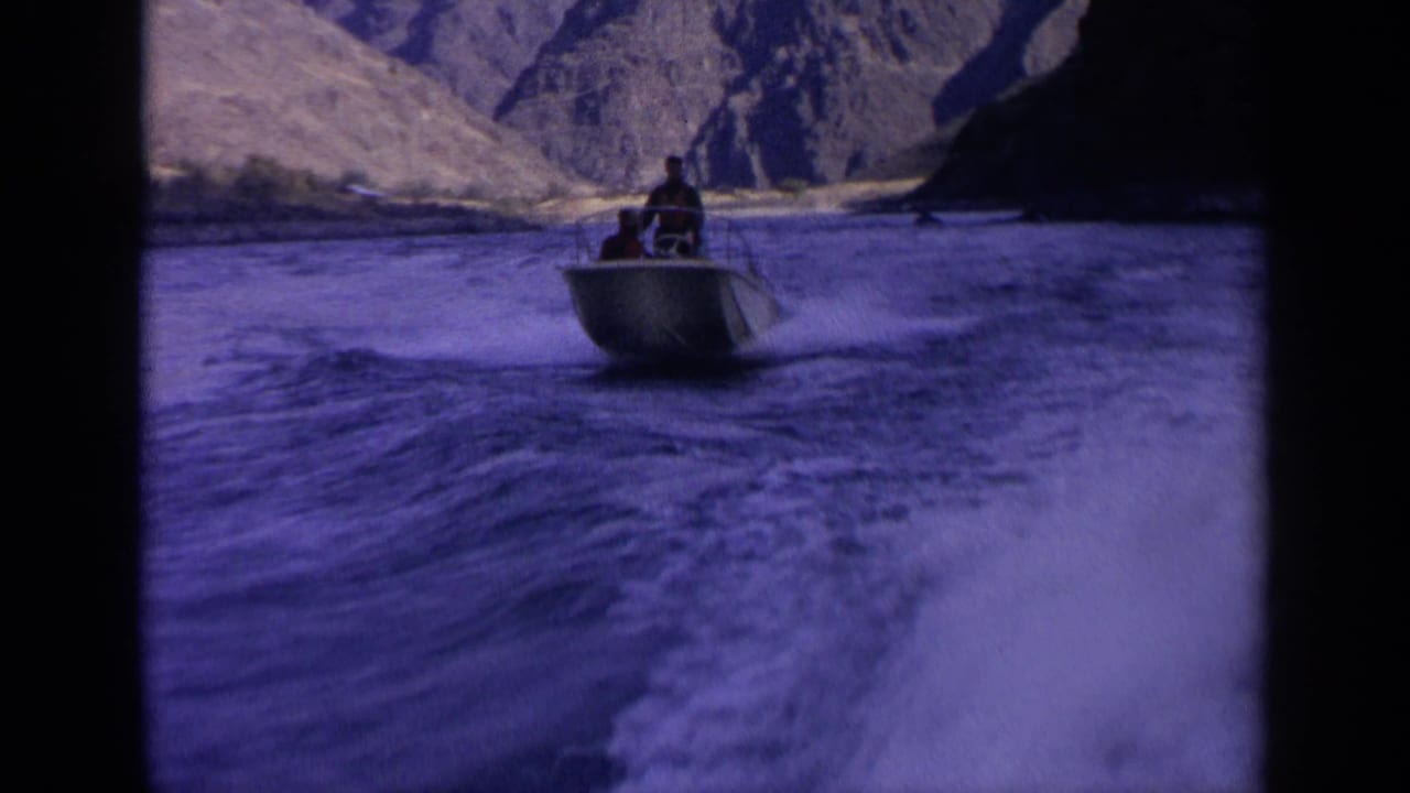 Small boat with two men continues down an Idaho canyon river in 1963 home-movie footage, with foamy wake in the foreground.