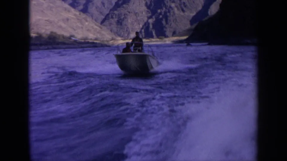 Small boat with two men continues down an Idaho canyon river in 1963 home-movie footage, with foamy wake in the foreground.