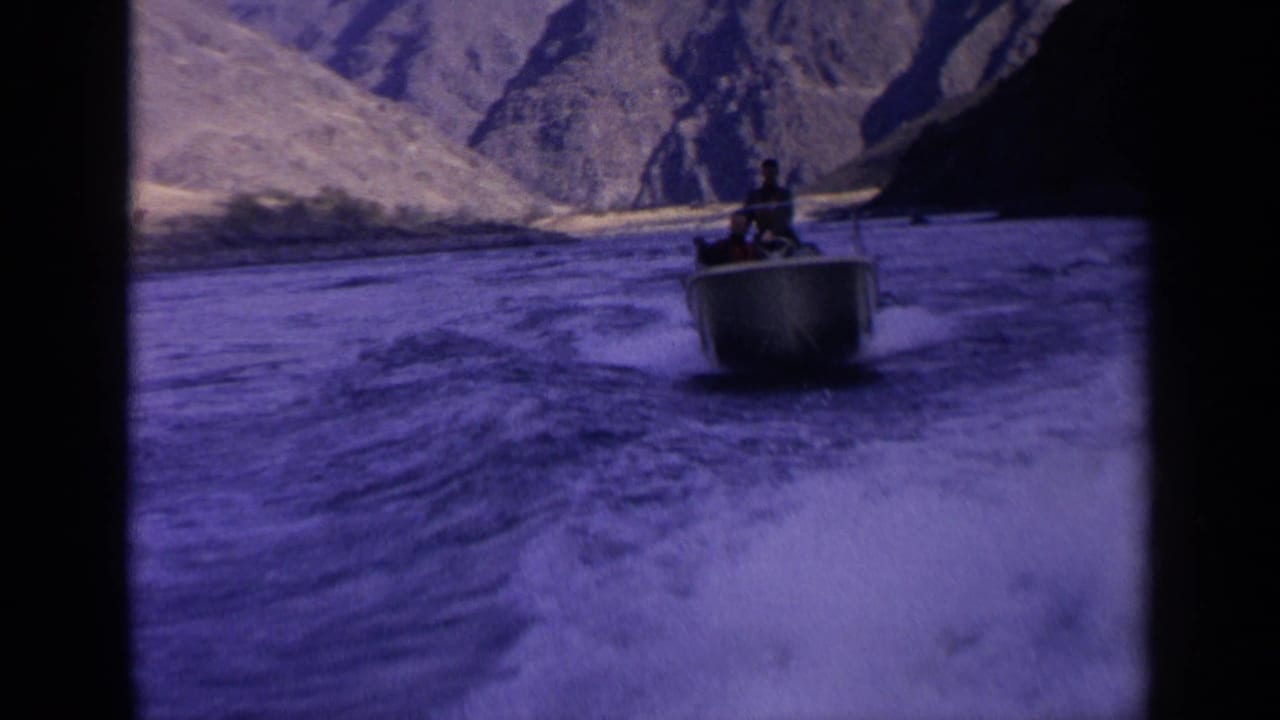 A motorboat carrying distant passengers moves across turbulent Idaho river water, framed by dry mountain slopes and aged film color shift.