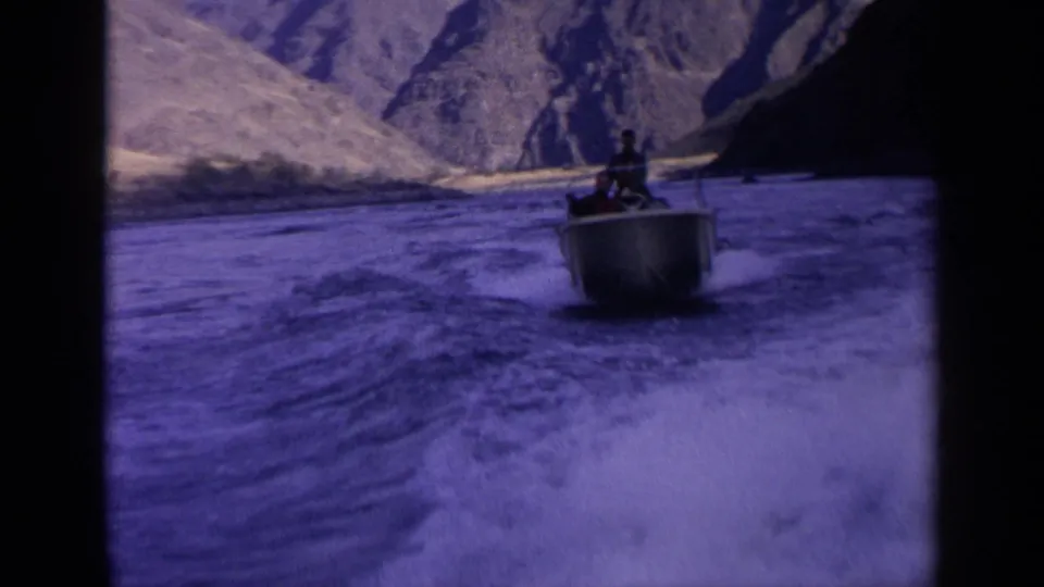 A motorboat carrying distant passengers moves across turbulent Idaho river water, framed by dry mountain slopes and aged film color shift.