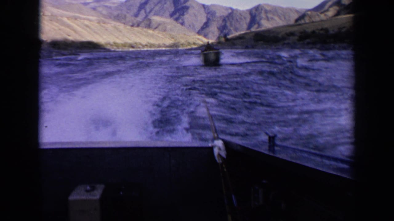 Moving-boat perspective on an Idaho river in 1963, with foamy wake, distant boat, and dry mountains under a bright sky.