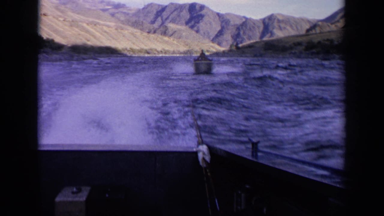 Home-movie view from a boat on an Idaho river, showing a distant craft, foamy wake, foreground deck, and rugged hills.
