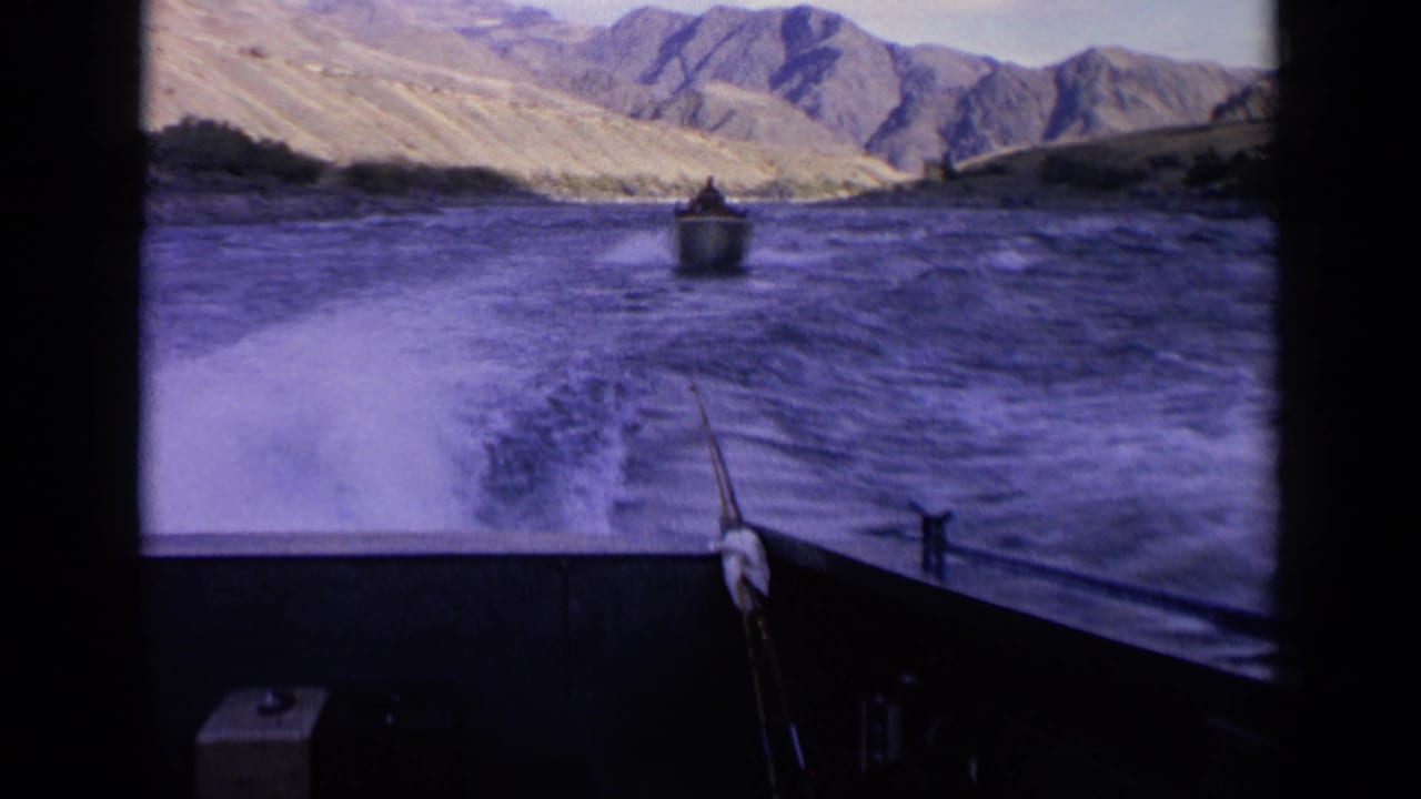 1963 Idaho river boating frame with foreground deck and rod, wake across the water, and a small boat moving toward mountains.
