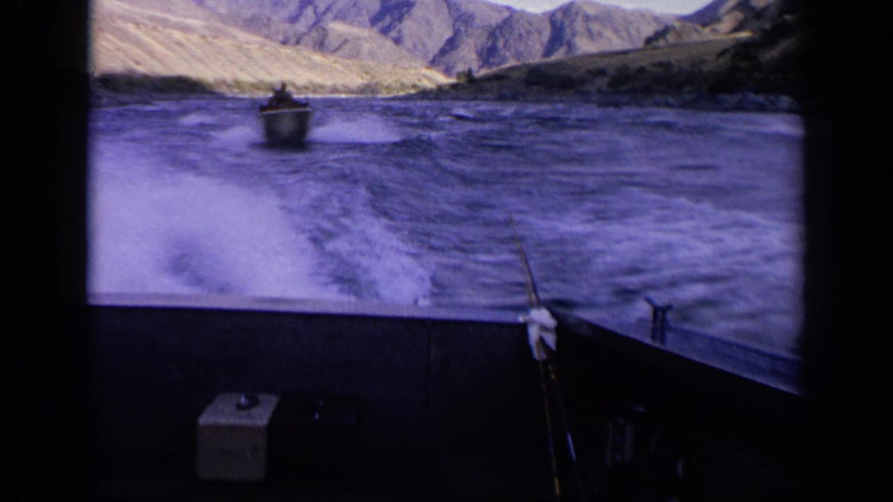Foamy wake behind the camera boat with a small boat ahead on a mountain-lined river.
