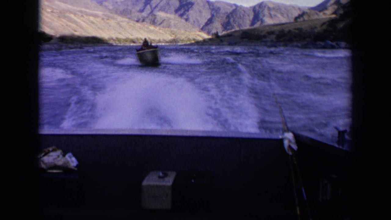 1963 Idaho 8mm boating view with a small boat crossing ahead, white wake behind the filming boat, and dry mountain terrain in the distance.