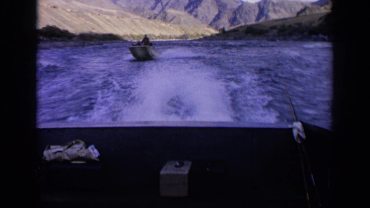 1963 Idaho home-movie frame from a moving boat, showing wake spray, a small boat with a person aboard, and sunlit mountain slopes.