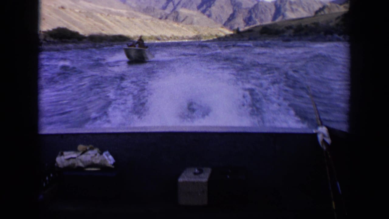 1963 Idaho 8mm view from a boat, looking past wake toward a small motorboat on a desert lake with rocky hills in the distance.