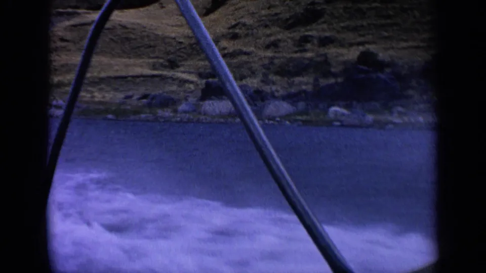 1963 Idaho home-movie frame from a moving boat, showing a fishing rod, foamy water, and dry shoreline along the lake.