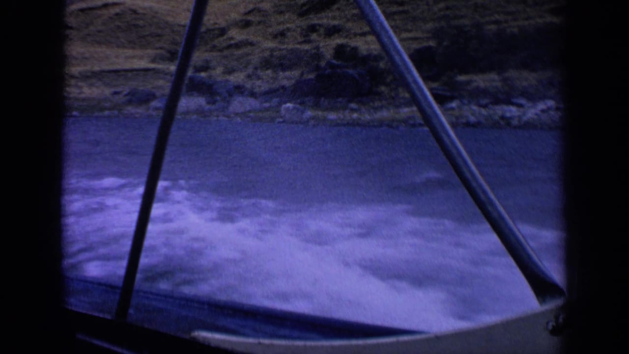 1963 home-movie frame looking out from a moving boat, showing churning wake, dark railings, blue water, and distant rocks and grass.