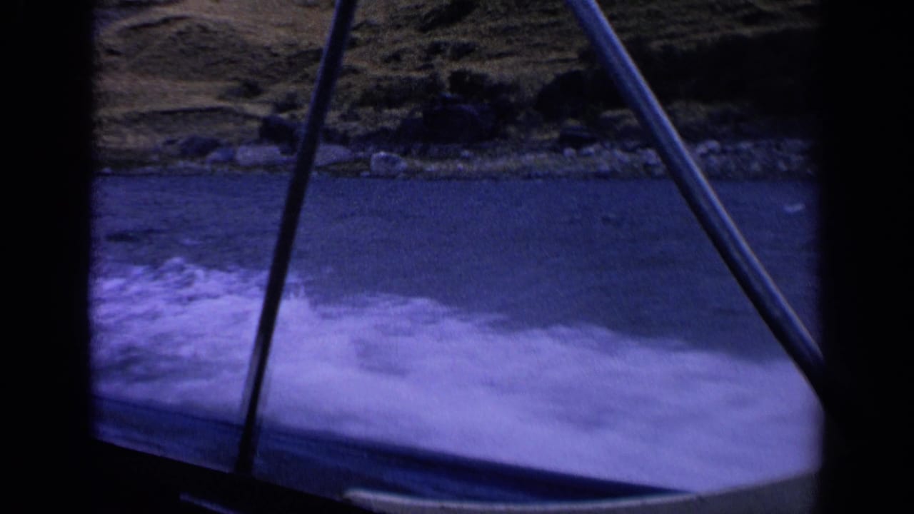 Final 8mm frame from the moving boat view in Idaho, with crossed metal railings, white wake, blue water, and a rocky grassy shoreline.