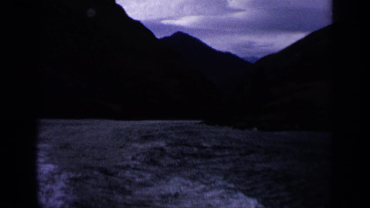 1963 Idaho lake view from a moving boat, with dark mountain ridges and purple overcast sky visible through the vessel structure.