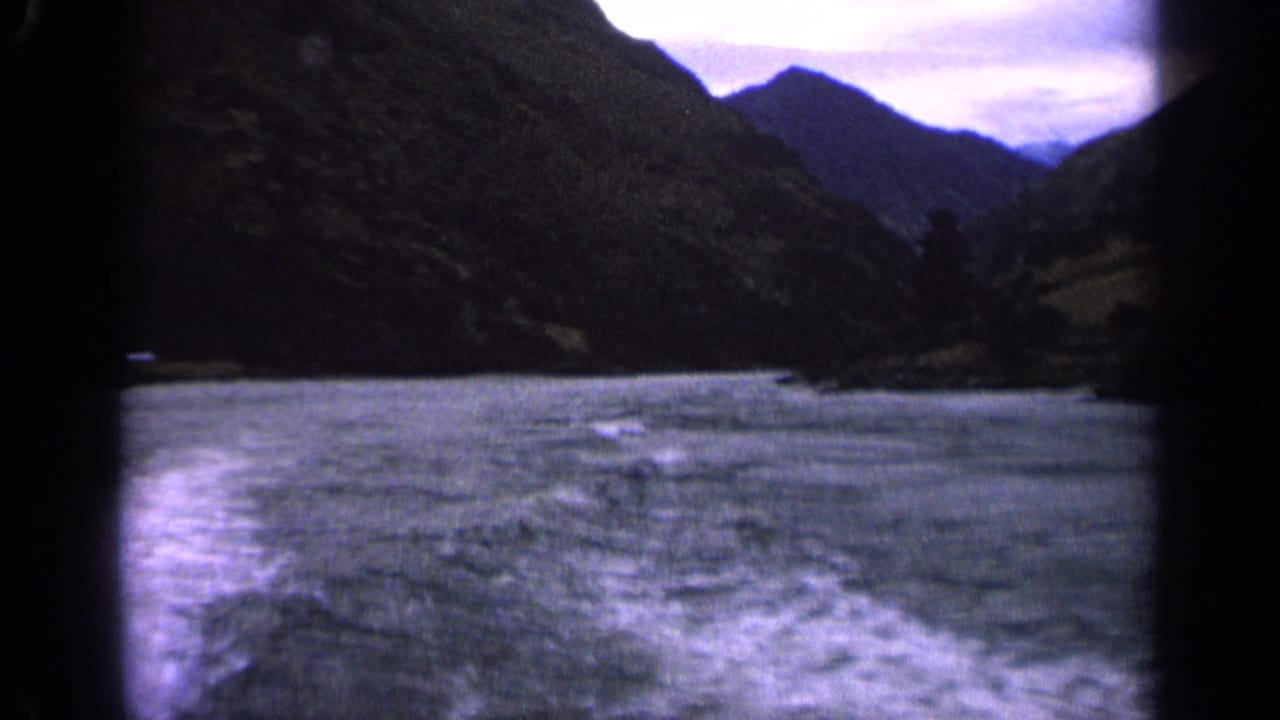 1963 Idaho home-movie frame with rippled lake water, white wake, and dark mountain slopes in a blue-purple film cast.