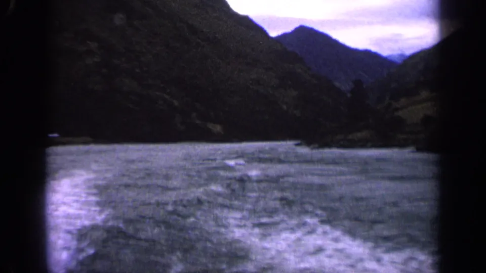 1963 Idaho home-movie frame with rippled lake water, white wake, and dark mountain slopes in a blue-purple film cast.