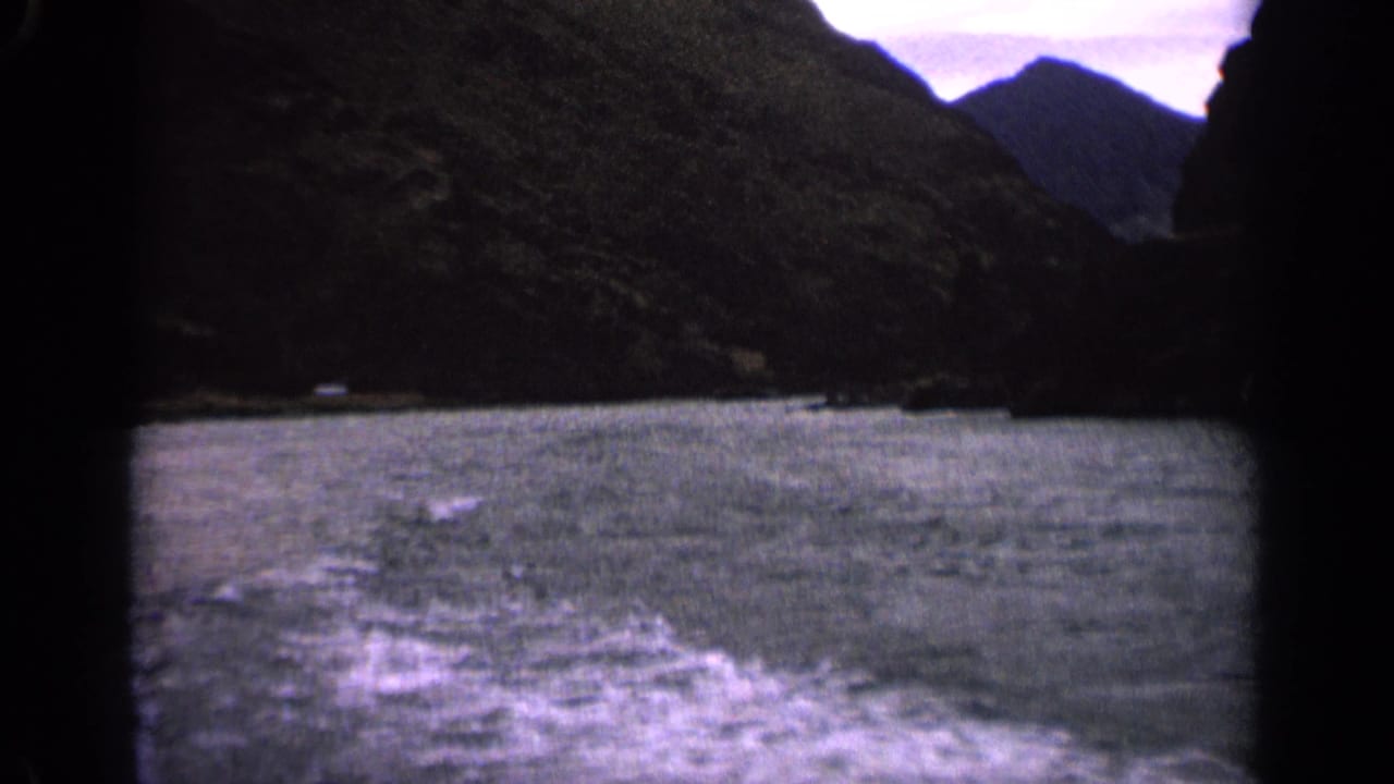 1963 Idaho home-movie frame with foaming wake on choppy water, rugged mountain slopes, and a muted cloudy sky.