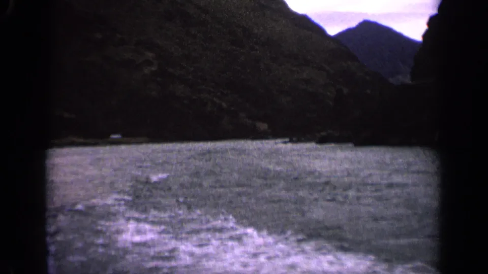 1963 Idaho home-movie frame with foaming wake on choppy water, rugged mountain slopes, and a muted cloudy sky.