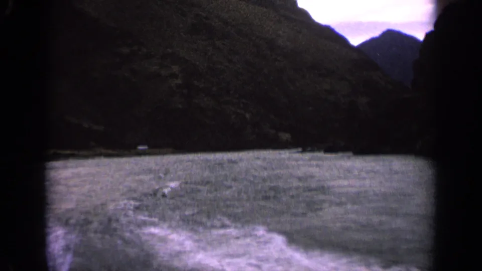1963 Idaho 8mm frame looking across lake water from a moving boat toward dark mountain shoreline and pale sky.