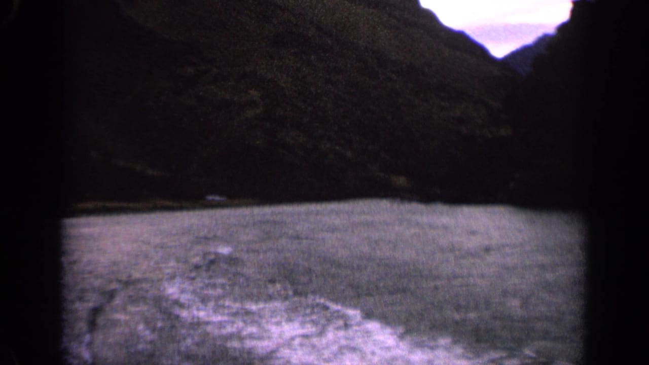 1963 Idaho home-movie frame showing a boat wake spreading across lake water below a dark mountain shoreline.