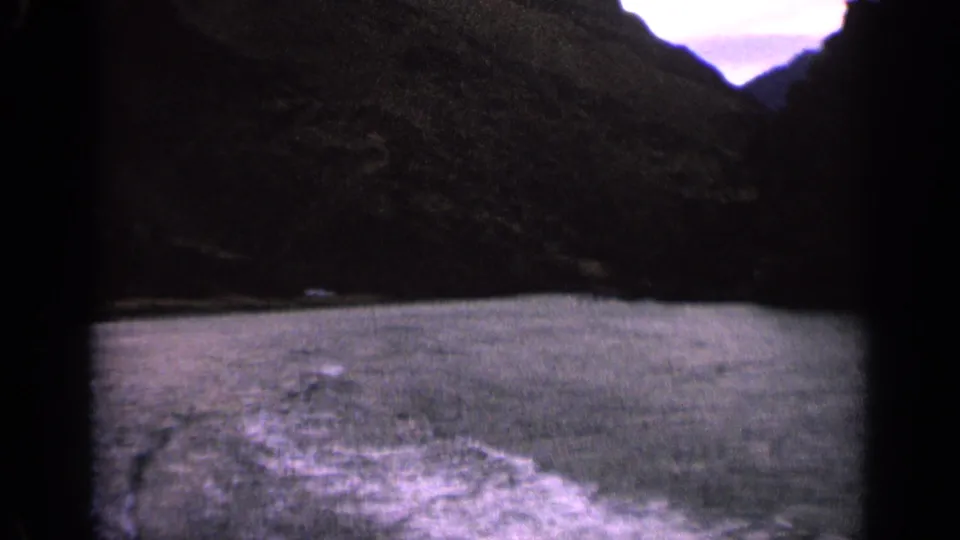 1963 Idaho home-movie frame showing a boat wake spreading across lake water below a dark mountain shoreline.