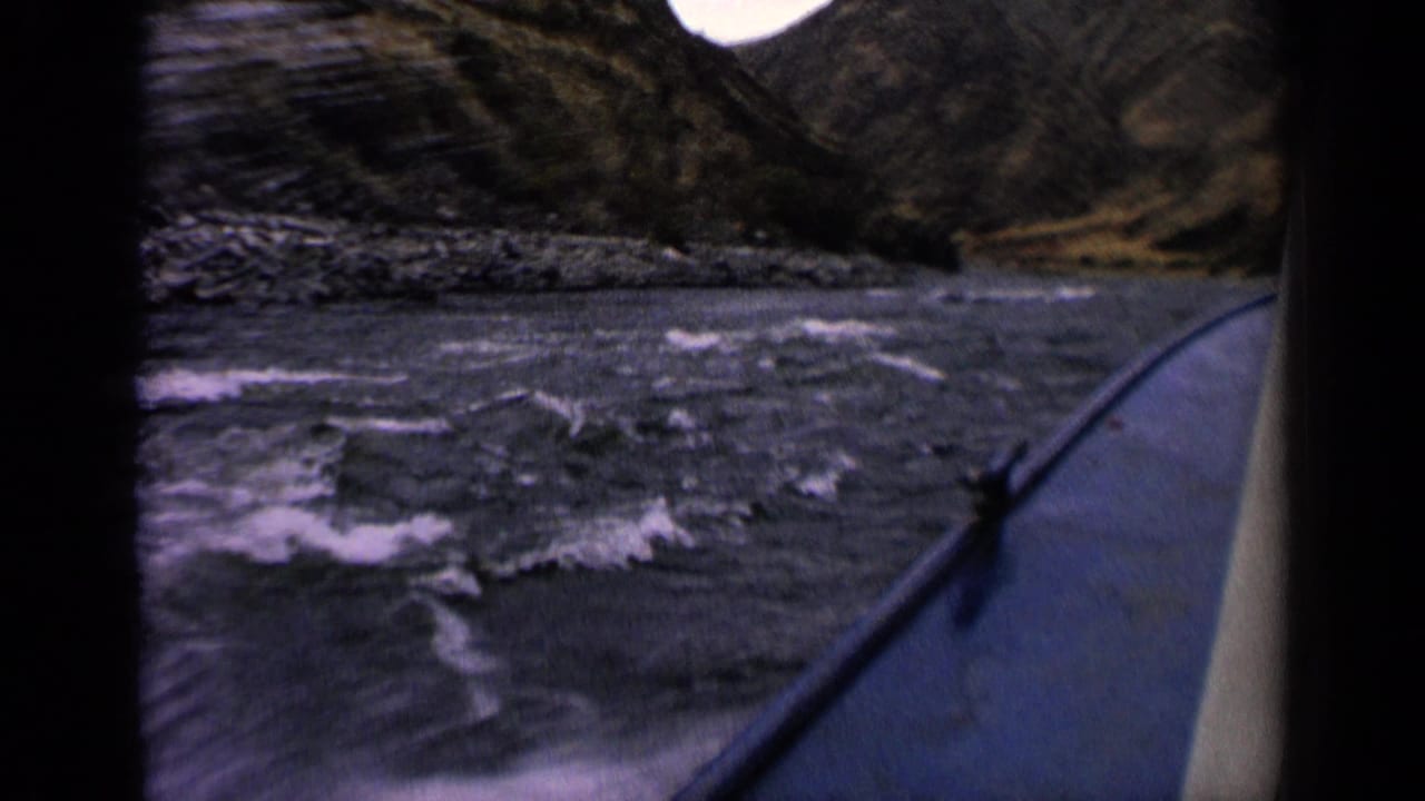1963 Idaho river view from a boat, with a rail at right, rough water ahead, and rocky banks along the channel.