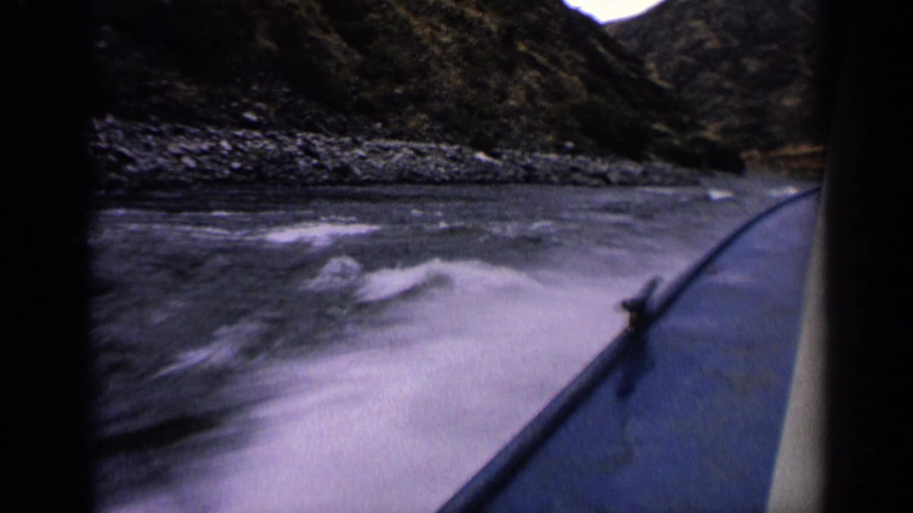 Moving-boat perspective on a rocky Idaho river, with foamy wake, boat side, and steep banks visible in the 8mm frame.