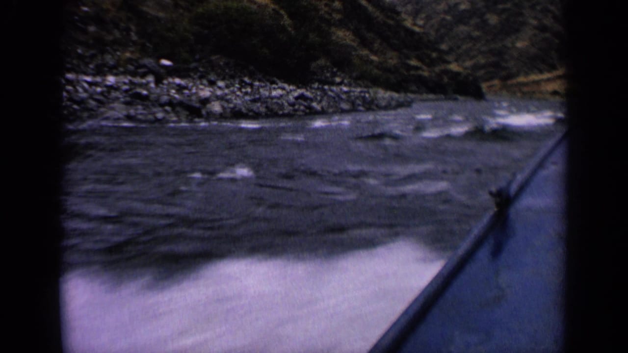 First-person 8mm view from a boat on an Idaho river, with white ripples, rocks, and tree-covered slopes visible ahead.