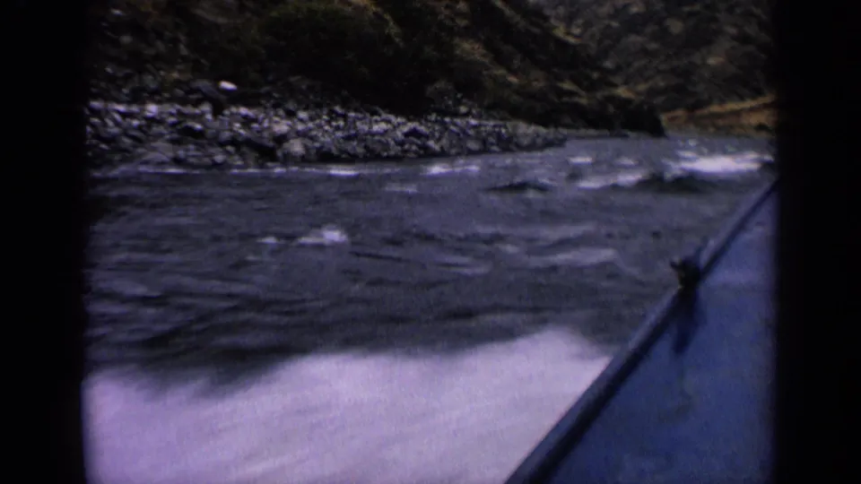First-person 8mm view from a boat on an Idaho river, with white ripples, rocks, and tree-covered slopes visible ahead.