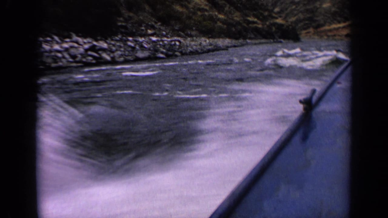 A 1963 home-movie frame from a boat shows churning river water, a blue rail, and a rugged shoreline in Idaho.