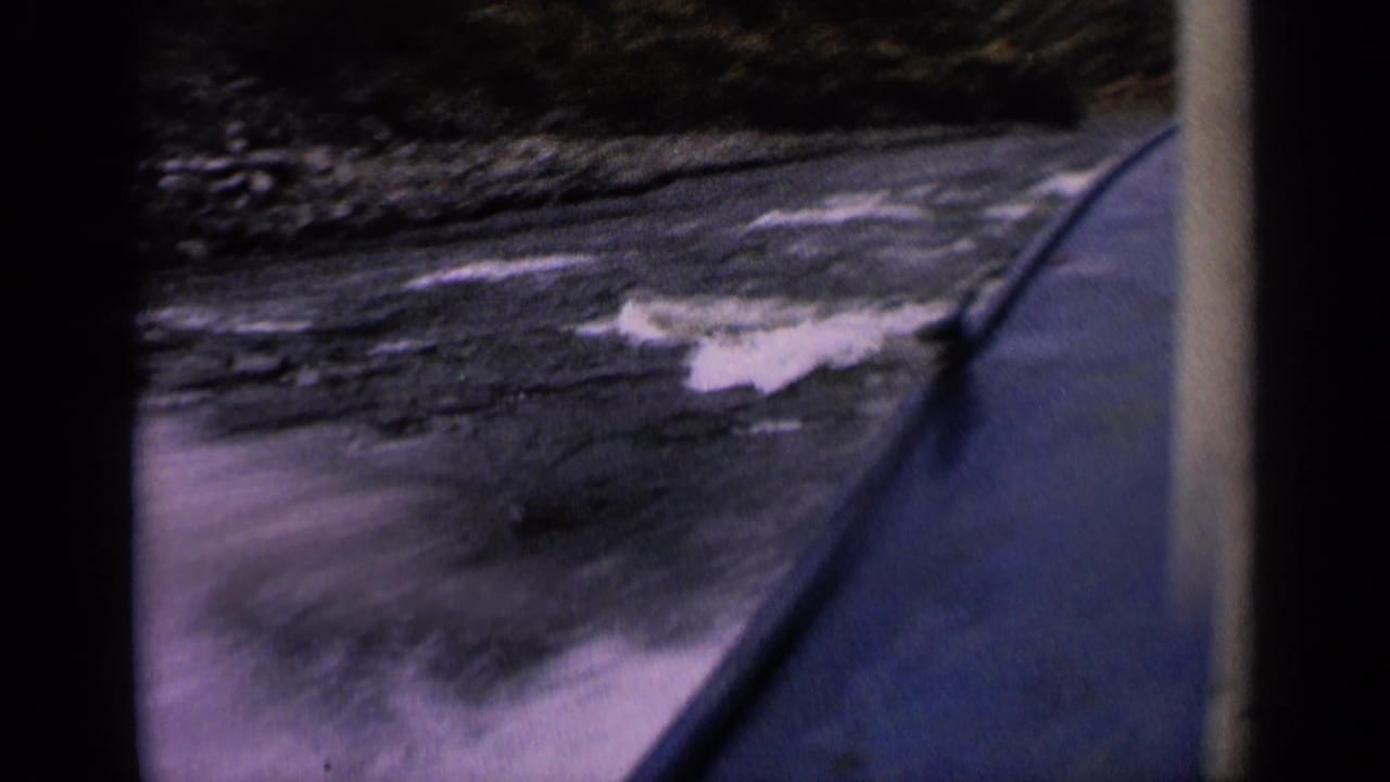 Vintage 8mm boat-level view of small rapids and rocky riverbank, with purple tint and dark frame edges from the film scan.