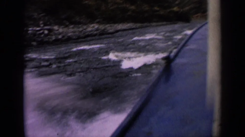 Vintage 8mm boat-level view of small rapids and rocky riverbank, with purple tint and dark frame edges from the film scan.