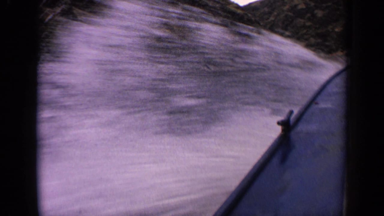 Handheld 1963 lake POV with the speedboat bow at right and blurred water below a steep Idaho mountainside.
