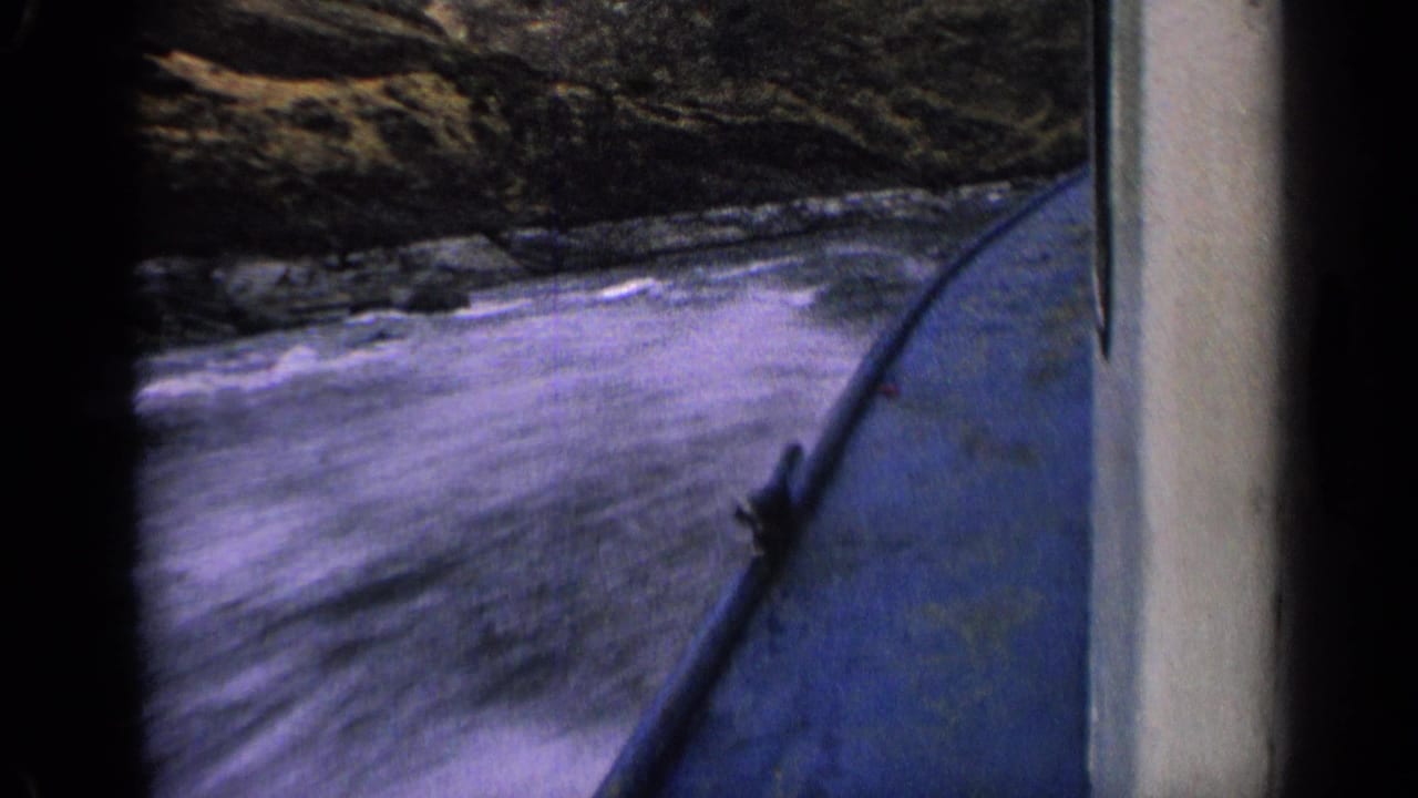Blue boat side cutting through splashing river water beside a rocky bank.