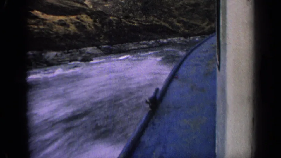 1963 Idaho 8mm frame showing the side of a blue boat as river water splashes against the hull near a rocky shoreline.