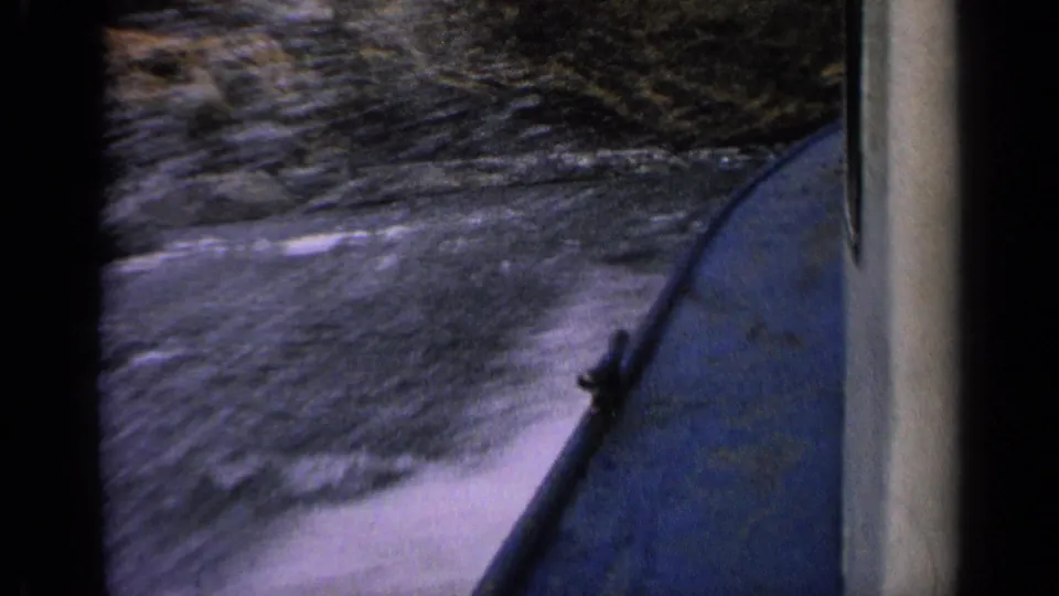 1963 Idaho home-movie frame with the blue boat side at right and blurred water rushing past a rocky bank.