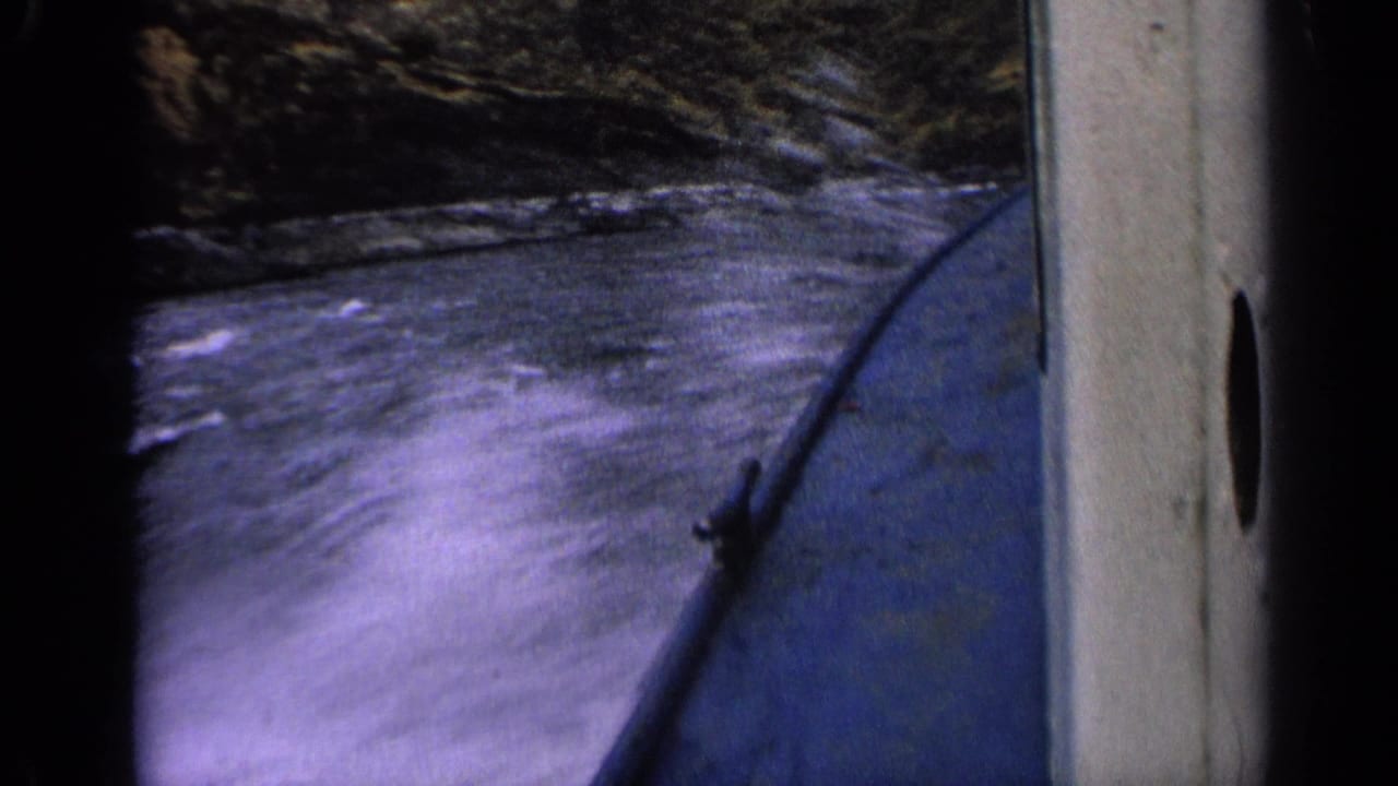 1963 Idaho 8mm frame looking down at foaming river water beside a blue boat, with dark rocky shoreline visible above.