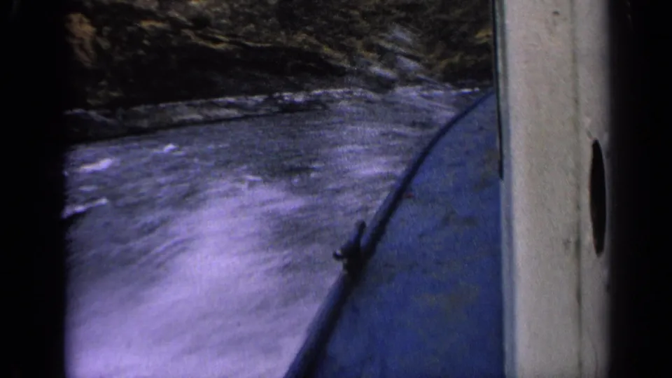 1963 Idaho 8mm frame looking down at foaming river water beside a blue boat, with dark rocky shoreline visible above.