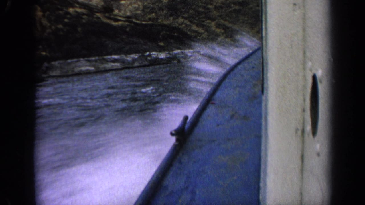1963 Idaho home-movie frame showing spray along the side of a blue boat as it passes rocky river terrain.