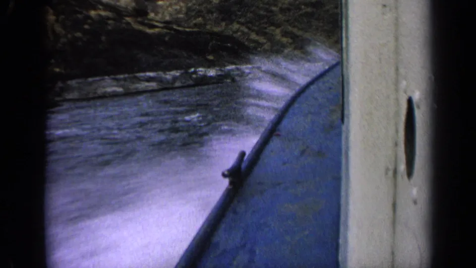 1963 Idaho home-movie frame showing spray along the side of a blue boat as it passes rocky river terrain.