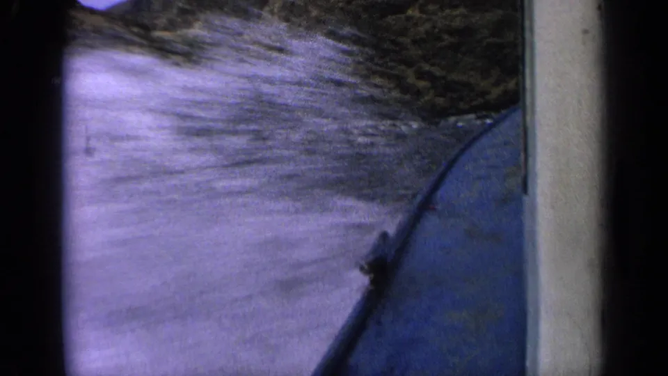 1963 Idaho 8mm frame showing a blue boat hull at speed with splashing river water and a shadowed shoreline nearby.