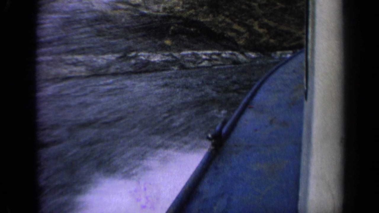 First-person 1963 Idaho boating view with the blue hull at right, rippled water below and rocks along the bank.