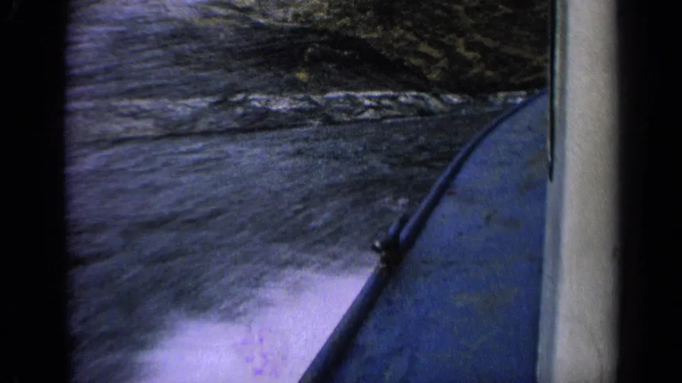 First-person 1963 Idaho boating view with the blue hull at right, rippled water below and rocks along the bank.