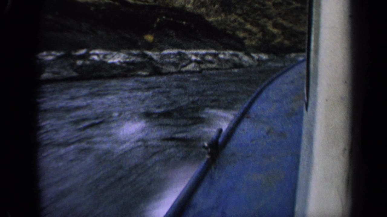 Handheld 8mm boating frame from Idaho in 1963, showing river wake, a blue hull and a rocky shoreline in motion.