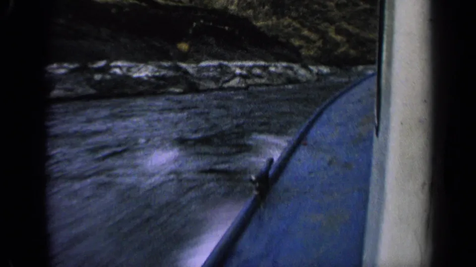 Handheld 8mm boating frame from Idaho in 1963, showing river wake, a blue hull and a rocky shoreline in motion.
