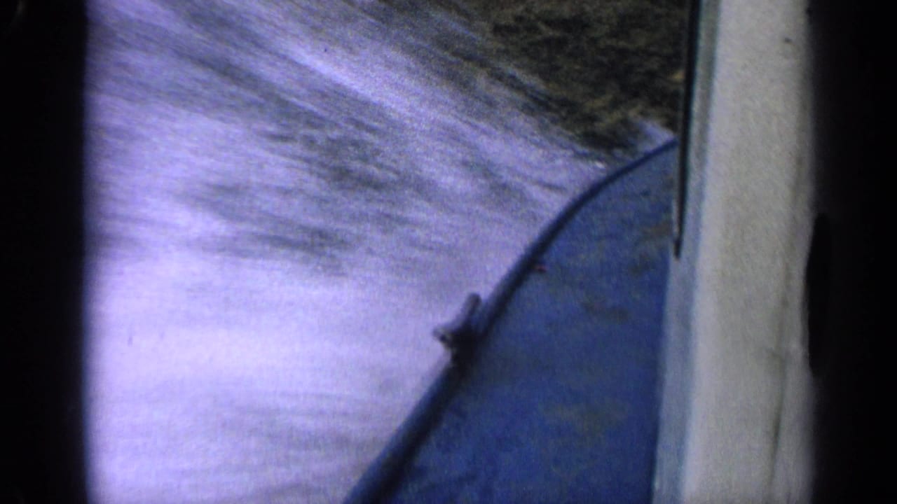 1963 Idaho home-movie frame with blue boat hull at right and bright, motion-blurred water splashing alongside.